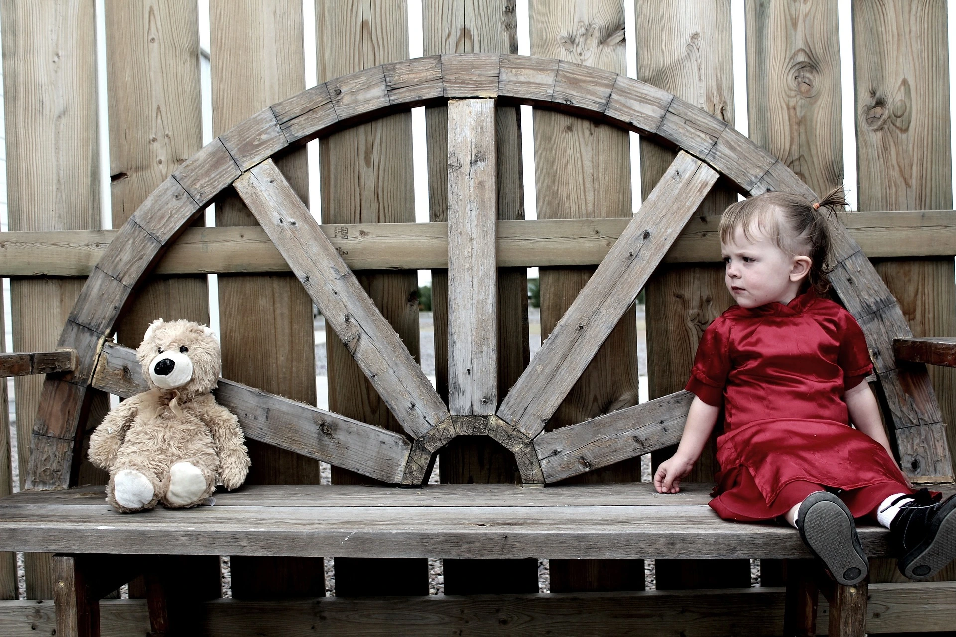 O fetita îmbrăcată intr-o rochie roșie, sta pe o banca la o distanta mică de un ursuleț de plus uitându-se spre el.