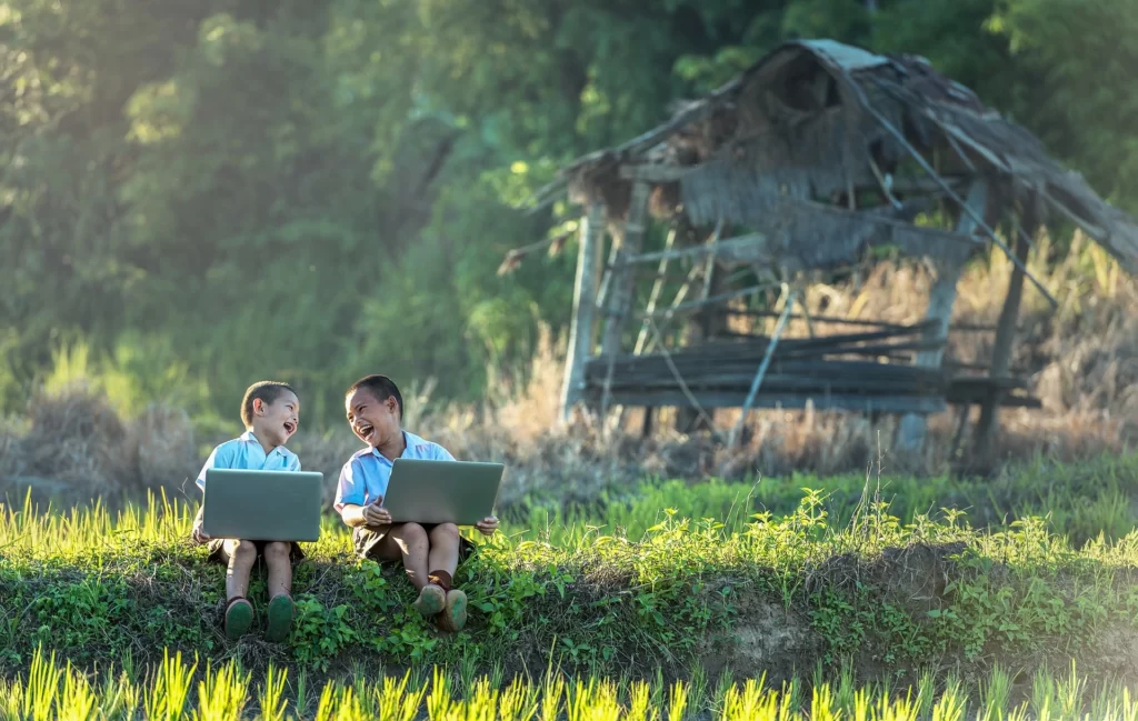 Doi băieți stau pe iarba cu laptopurile lor, râzând împreună.